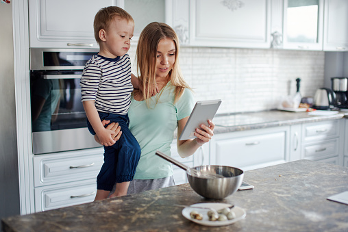 Portrait of young blond mother holding toddler son at the kitchen. She is looking at tablet screen, cooking something