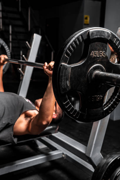 fotografía de alta calidad. hombre hispano con camisa negra en un gimnasio acostado levantando un peso pesado con ambos brazos. 45 libras de peso siendo levantado. - arms lifted fotografías e imágenes de stock