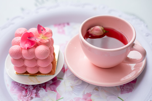 Serving festive tray of appetizing creamy cake with butterfly decoration and pink cup of fragrant tasty tea with flower petals top view closeup. Pink style decor, holiday, cute service.
