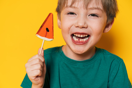 Boy in green t-shirt holding lollipop in the form of piece of watermelon in his hand against yellow background.