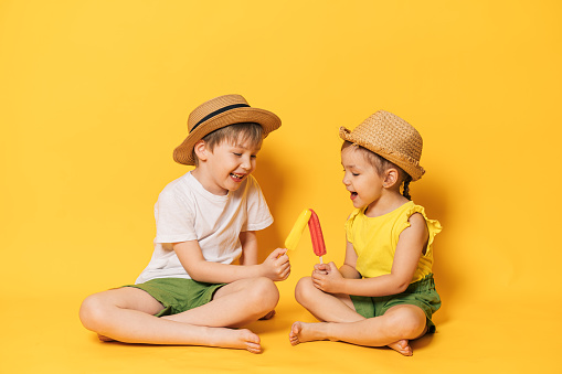 Children holding ice cream on stick sit on yellow background.
