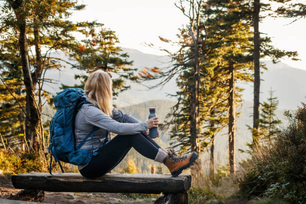 Woman with backpack and thermos sitting on bench in forest stock photo