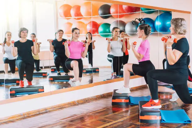 Group of women are engaged in step aerobics with dumbbells in their hands in front of mirror.