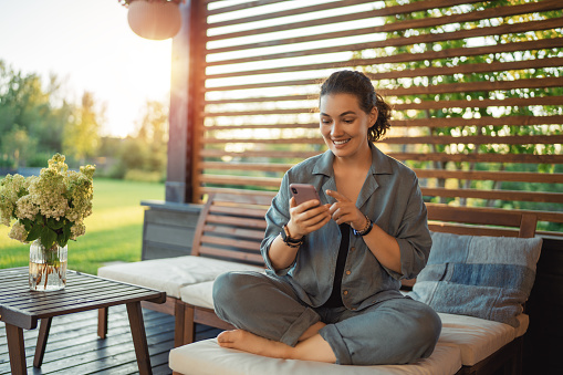 Happy young woman is using a phone sitting on the patio in summer.
