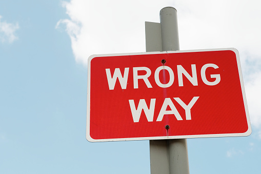 A red rectangular Wrong Way sign post against a blue sky with some clouds.  Advice to drivers or can be used as a metaphor for business and personal decisions