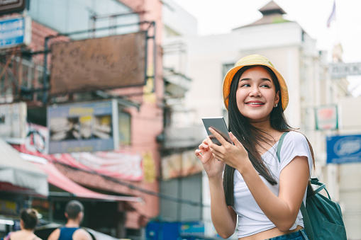 Travel vacation city concept, Young happy traveler asian woman with backpack using mobile phone in Khaosan Road outdoor market in Bangkok, Thailand