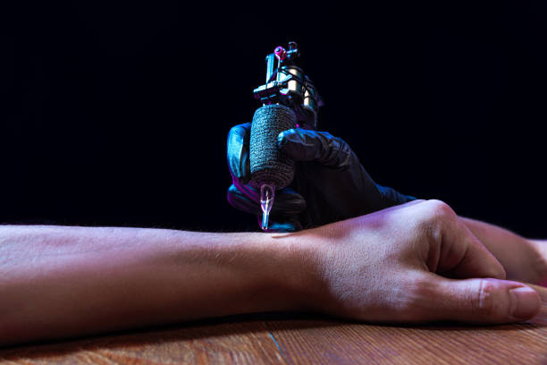 tattooer master's hand in black glove making tattoo art on male hand with machine for tattoo over dark background in neon light. beauty, style, fashion - dövme yaptırmak fotoğraflar stok fotoğraflar ve resimler