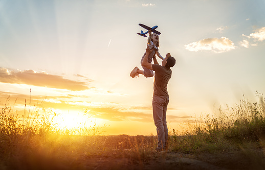 Happy family, father and kid with toy plane are playing on the field at sunset.