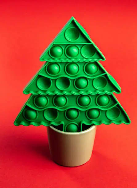 Photo of Green kids anti stress pop it toy as Xmas tree standing in beige flower pot on red backdrop.