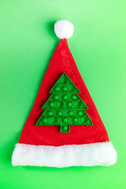 Photo of Trendy popular child anti stress pop it toy in form of green Xmas tree on red Santa's hat on green.