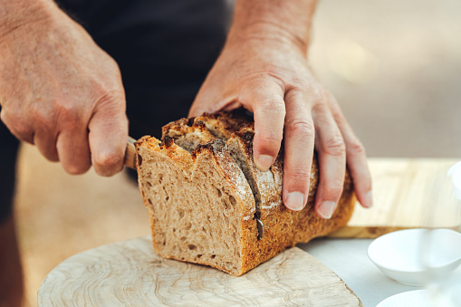 Close-up of senior man hand cutting slice from loaf of bread with kitchen knife in table in garden. male hands  cutting bread on table outdoors.