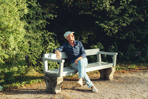 Senior man wearing cap sitting alone on bench in the park. Retired person soaking sunlight sitting on park bench on a summer day.
