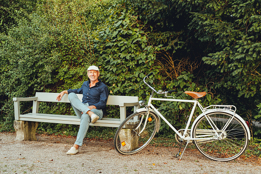 Senior man wearing cap sitting alone on bench in the park with his bicycle parked by. Mature person sitting on park bench and smiling.