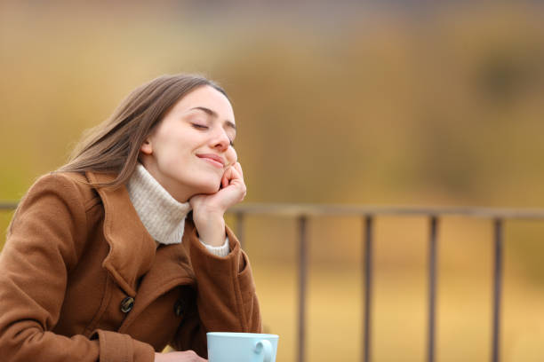 A woman dressed warmly in a winter coat enjoys a hot beverage outside of a house  on a snowy day, surrounded by trees and winter scenery