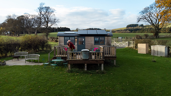 A shot of luxury wooden huts overlooking a grass field at a glamping site in Alnwick, Northumberland. One senior man is standing on the decking relaxing.