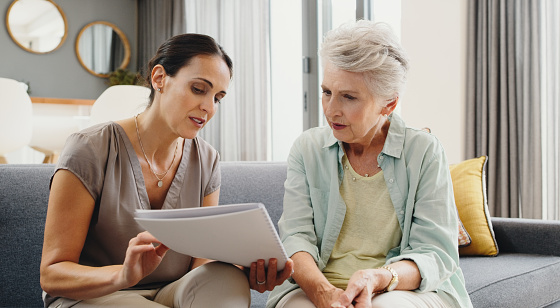 Caregiver talking to senior woman about insurance, support with finance documents and help reading retirement paperwork on the sofa in home. Girl helping elderly person with Alzheimer care at house