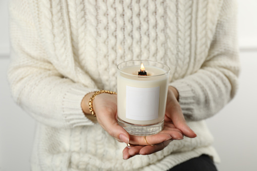 Woman with stylish jewelry holding burning soy candle on light background, closeup