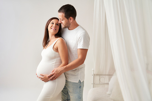 Beautiful pregnant woman and her handsome husband smile happily as they look at each other while embracing
