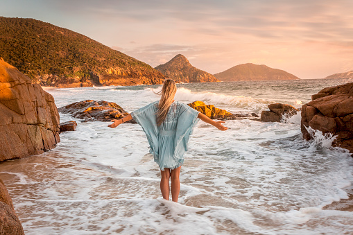 Woman wearing a flowing blue dress, stands in ocean waves and looks out to distant volcanic mountains and  soft pastel sunrise sky