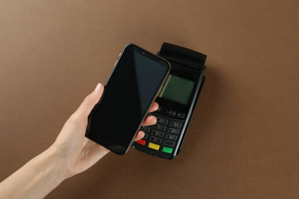 istock Woman with smartphone using modern payment terminal on brown background, top view 1423023492