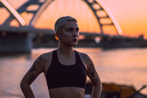 Smiling young tattooed woman with short hair on an early morning outside workout. Determined and disciplined young woman out for a run, during a beautiful sunrise by the river and a bridge.