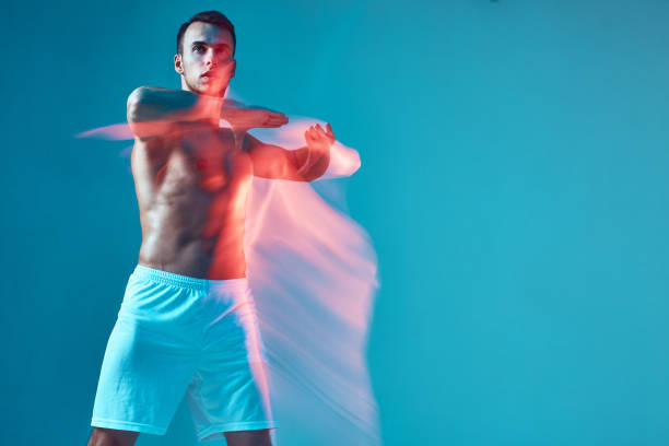 istock Athletic guy with naked torso raised arms performing dance on blue studio background. Copy space. Long exposure 1423032175
