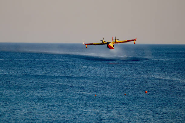 Red and yellow rescue plane leaves spray of seawater behind when scooping up seawater. Small red buoys in the foreground contrasting with the blue sea.