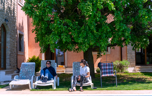 Two mature woman talking while sitting on deck chair in front of the swimming pool in house backyard. That is in small place Peroj near Pula in Istria, Croatia with Brioni islands near by.
