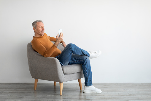 People And Technology Concept. Portrait of casual cool mature man resting and using smart phone sitting on chair isolated on white studio background wall, free copy space mockup, full body length