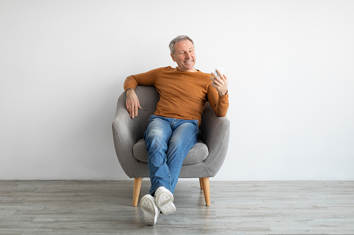 Portrait of smiling mature man using mobile phone, watching video sitting resting on armchair isolated at white background wall. Guy browsing internet, surfing web, free copy space, full body length