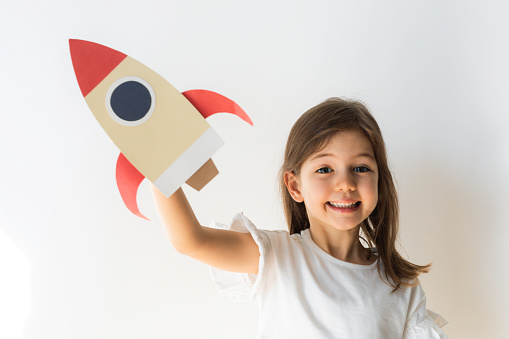 Little girl  is holding model rocket in hand and is smiling  in front of white background.