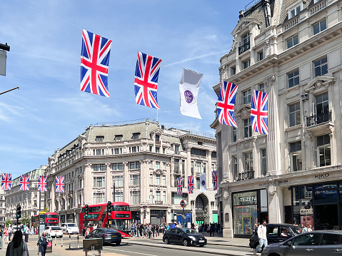 Oxford Street in downtown London decorated with Union Flags and the official logo for the Platinum Jubilee of Queen Elizabeth II in May 2022. Red double decker buses along with taxis and people fill the street in a sunny day. Many garlands with Union Jack (British flag).