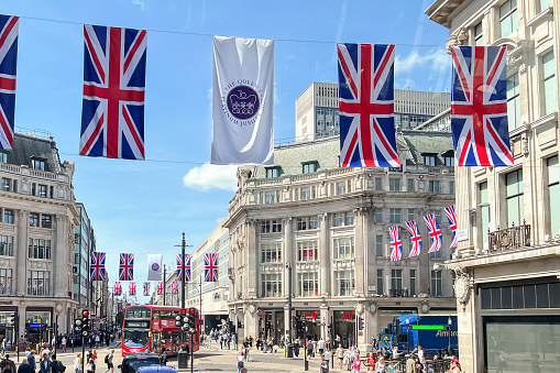 Oxford Circus in downtown London decorated with British flags and the official logo for the Platinum Jubilee of Queen Elizabeth II in May 2022. Sunny day, with buses, taxis and pedestrians filling the streets.