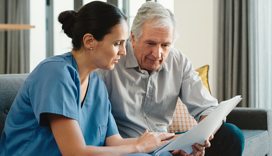 A doctor, nurse or medical employee consulting senior man on a sofa in a hospital lounge. Woman healthcare worker going over insurance paperwork, chart or diagnosis with a male patient in retirement.