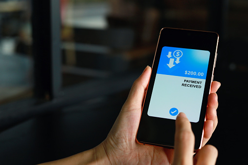 Cropped image of a woman's hand holding smartphone, receiving money through digital wallet from a friend in a cafe. Smart banking with technology.