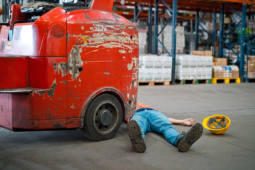 Man lying down on floor in front of in forklift in a warehouse.