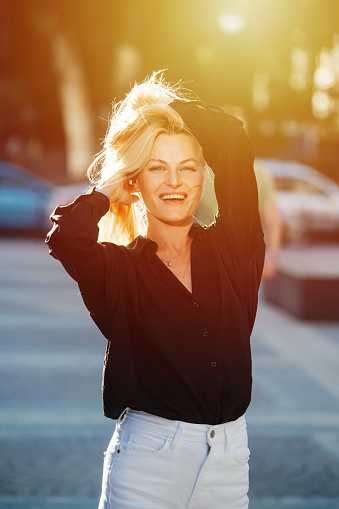 Radiant attractive young blond woman ruffling her hair looking at the camera. She is standing on a pavement on a sunlit street, against blurred background.