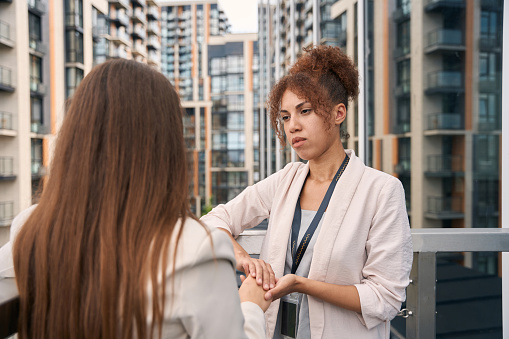 Compassionate corporate worker standing on office balcony and holding her female colleague by hand
