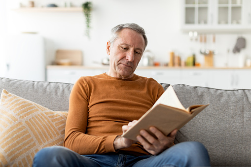 Leisure and Relaxation Concept. Portrait of casual mature man holding paper book or photo album, sitting on couch at home in living room, reading literature, enjoying story, free copy space