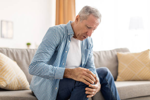 Mature man with knee pain sitting on couch at home Trauma And Rheumatoid Arthritis Concept. Sad mature male holding touching his knee feeling acute pain, sick senior guy suffering from aching leg sitting on couch at home, selective focus on hand osteoarthritis stock pictures, royalty-free photos & images