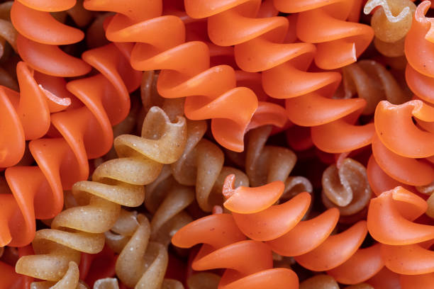 A detailed close-up of dried raw lentil rotini pasta, showcasing its spiral shape and vibrant orange hue. The tightly packed arrangement highlights the texture and structure of the pasta, emphasizing its plant-based composition. The image captures the intricate curves and edges of the pasta, revealing slight imperfections and breakages.