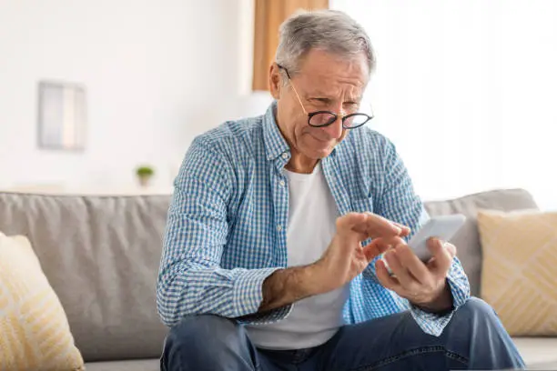 Poor Eyesight In Older Age, Macular Degeneration Concept. Confused Mature Male In Eyeglasses Squinting Eyes Reading Message On Cell Phone Having Ophtalmic Issue Problems With Vision Sitting On Sofa