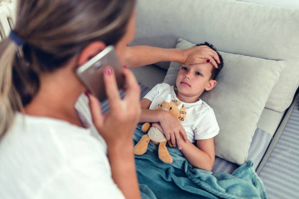 jeune mère blonde vérifiant la température de son garçon malade de 6 ans sur un canapé dans le salon de la maison, appelant le médecin. - 6 7 years photos et images de collection