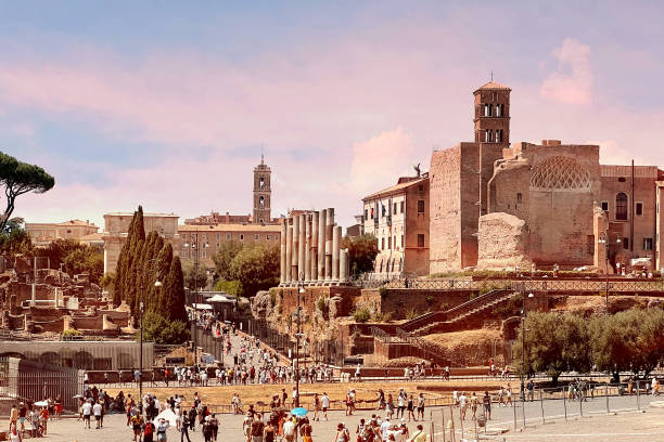 Plenty of tourists walking to the area of Roman Forum on a hot summer day, visiting monuments and archaeological ruins. The Capitol with its tower is visible at the end of the ancient road, framed by a colonnade.