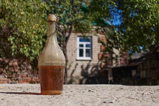 An old bottle of old sunflower oil lies on the ground, covered in dust and cobwebs. In the background there is a blue sky, green trees and a village house with a window. Selective focus.