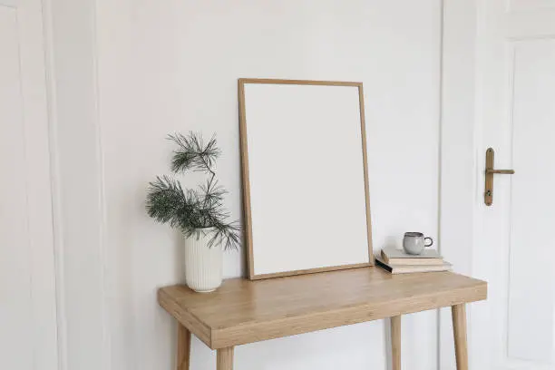 Photo of Christmas Scandinavian interior. Blank vertical wooden picture frame mockup. Pine tree branches in vase, cup of coffee and old books on table, desk. White wall background, doors. Empty copy space.