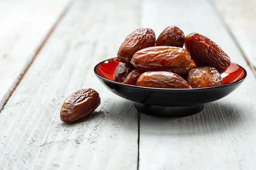 One Orange, black bowl with dried dates in white  wooden background. On the left of the bowl one date on the background.