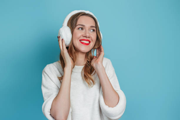 heureuse jeune femme caucasienne souriante avec des écouteurs en fourrure et un pull blanc souriant et regardant l’espace de copie isolé sur fond de studio bleu - ear muff photos et images de collection