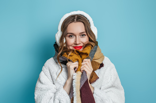 Portrait of young caucasian woman with red lips in white artificial eco fur coat isolated on blue studio background. Winter season.