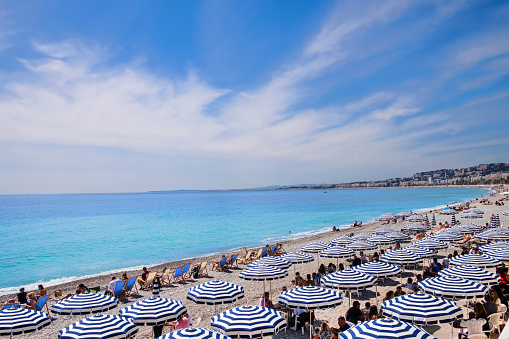 People sitting for the lunch under striped umbrellas on the long beach of the Promenade des Angles in Nice